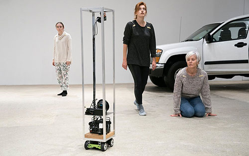 A minimalist robot stands in front of three actors and a pickup truck in a brightly-lit gallery during a play.